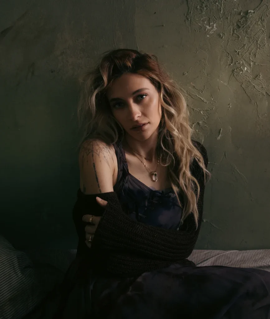 Paris Jackson with long wavy hair and shoulder tattoos sitting against a textured wall in dim lighting