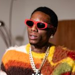 Soulja Boy wearing red sunglasses and a fuzzy multi-coloured sweater, holding two foam cups and a phone