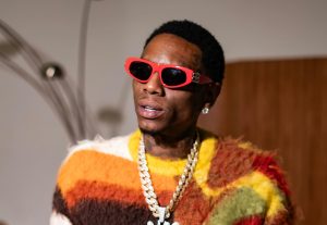 Soulja Boy wearing red sunglasses and a fuzzy multi-coloured sweater, holding two foam cups and a phone