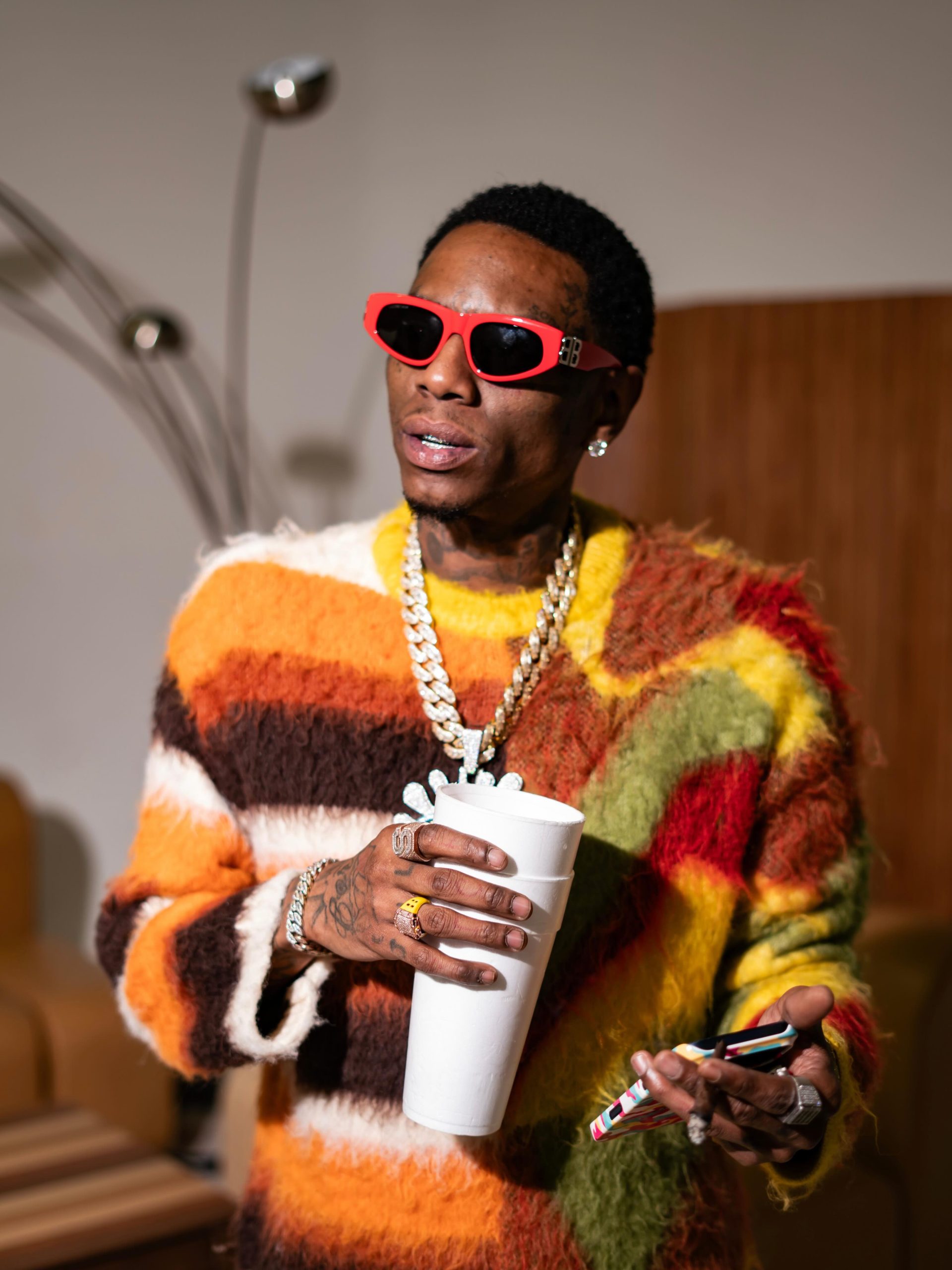 Soulja Boy wearing red sunglasses and a fuzzy multi-coloured sweater, holding two foam cups and a phone