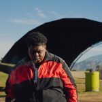 Bigxthaplug in a red and black Adidas jacket wearing a chain necklace looking down outdoors