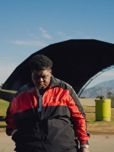 Bigxthaplug in a red and black Adidas jacket wearing a chain necklace looking down outdoors