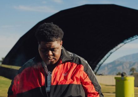 Bigxthaplug in a red and black Adidas jacket wearing a chain necklace looking down outdoors