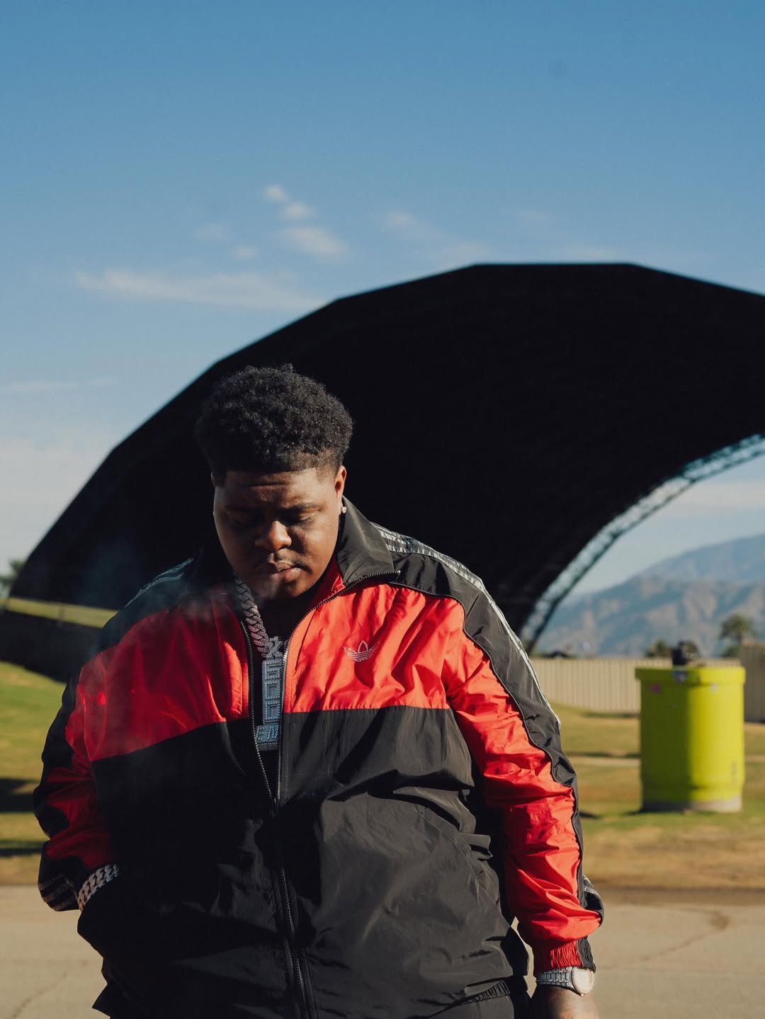 Bigxthaplug in a red and black Adidas jacket wearing a chain necklace looking down outdoors