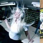 DJ_Dave wearing silver over-ear headphones with long blonde hair blowing wildly around her head in a workshop setting