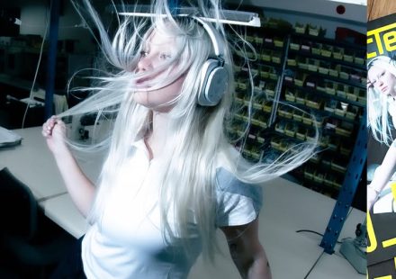 DJ_Dave wearing silver over-ear headphones with long blonde hair blowing wildly around her head in a workshop setting