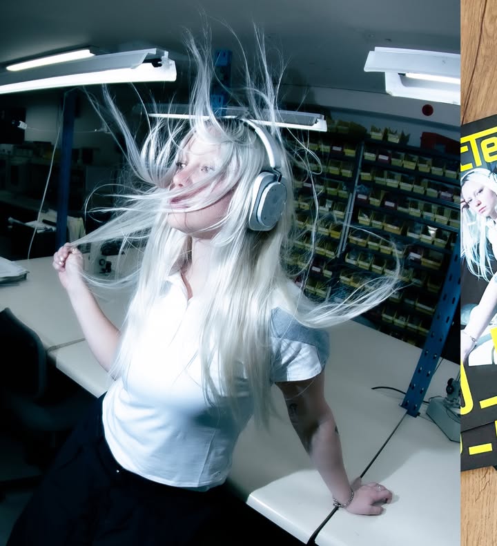 DJ_Dave wearing silver over-ear headphones with long blonde hair blowing wildly around her head in a workshop setting