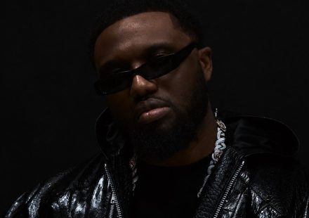 Headie One wearing dark rectangular sunglasses, a chain necklace, and a glossy black jacket against a black background