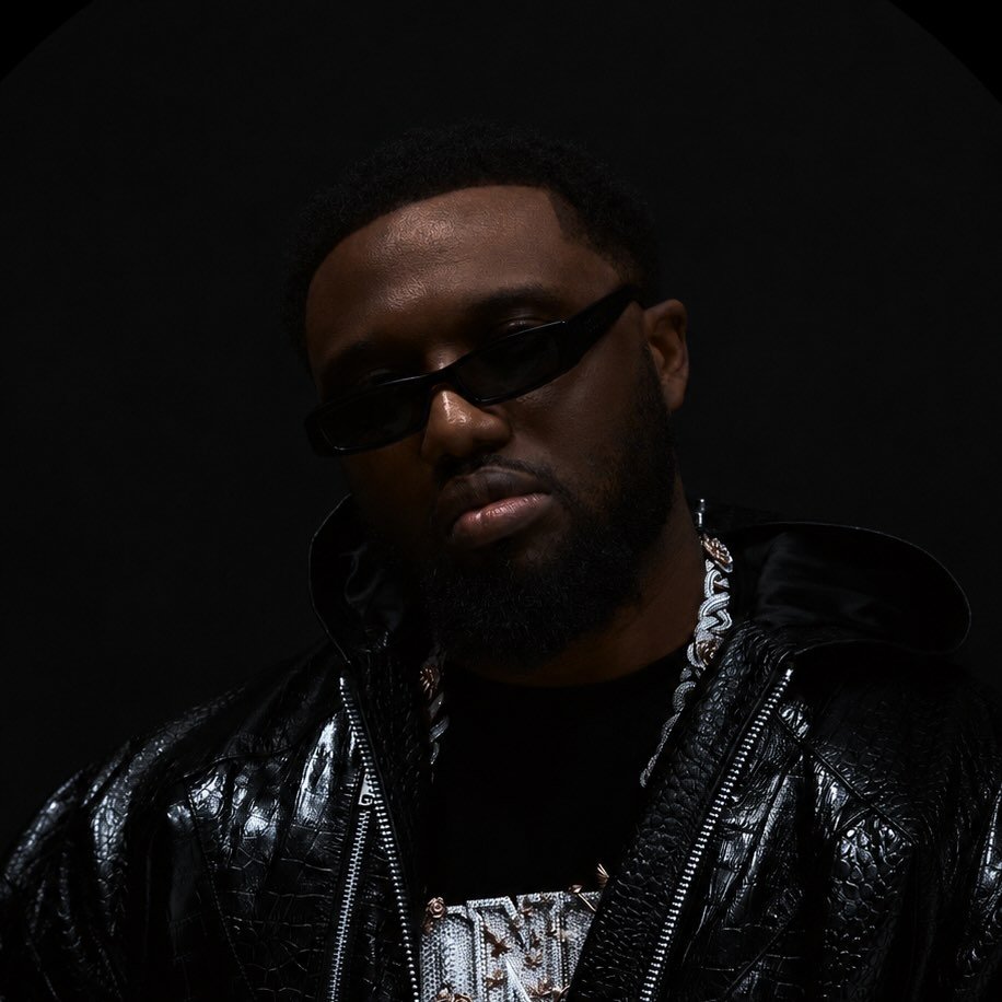Headie One wearing dark rectangular sunglasses, a chain necklace, and a glossy black jacket against a black background