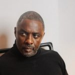 Idris Elba wearing a black turtleneck sweater whilst sitting in a chair and looking off-camera