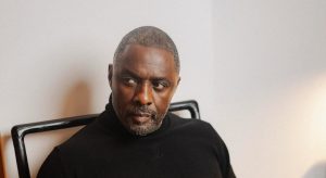 Idris Elba wearing a black turtleneck sweater whilst sitting in a chair and looking off-camera