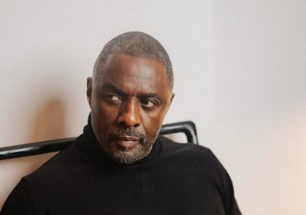 Idris Elba wearing a black turtleneck sweater whilst sitting in a chair and looking off-camera