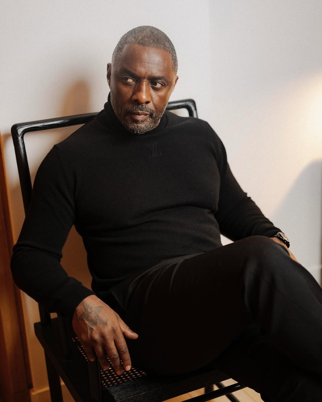 Idris Elba wearing a black turtleneck sweater whilst sitting in a chair and looking off-camera