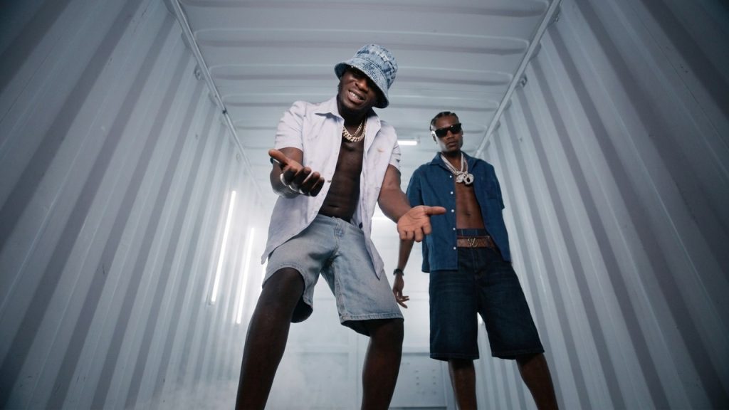 J Hus and Marnz Malone posing inside a lighted metal shipping container wearing unbuttoned shirts and chains