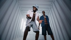 J Hus and Marnz Malone posing inside a lighted metal shipping container wearing unbuttoned shirts and chains