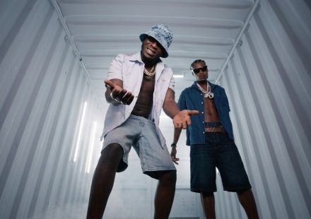 J Hus and Marnz Malone posing inside a lighted metal shipping container wearing unbuttoned shirts and chains