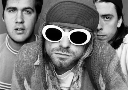 Kurt Cobain wearing white oval sunglasses and a trapper hat, flanked by Nirvana bandmates Krist Novoselic and Dave Grohl