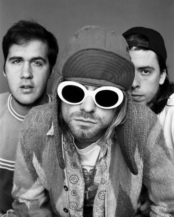 Kurt Cobain wearing white oval sunglasses and a trapper hat, flanked by Nirvana bandmates Krist Novoselic and Dave Grohl