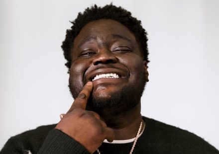 Swayvo Twain touching his lip whilst wearing a black sweater with white patterns and a gold chain necklace