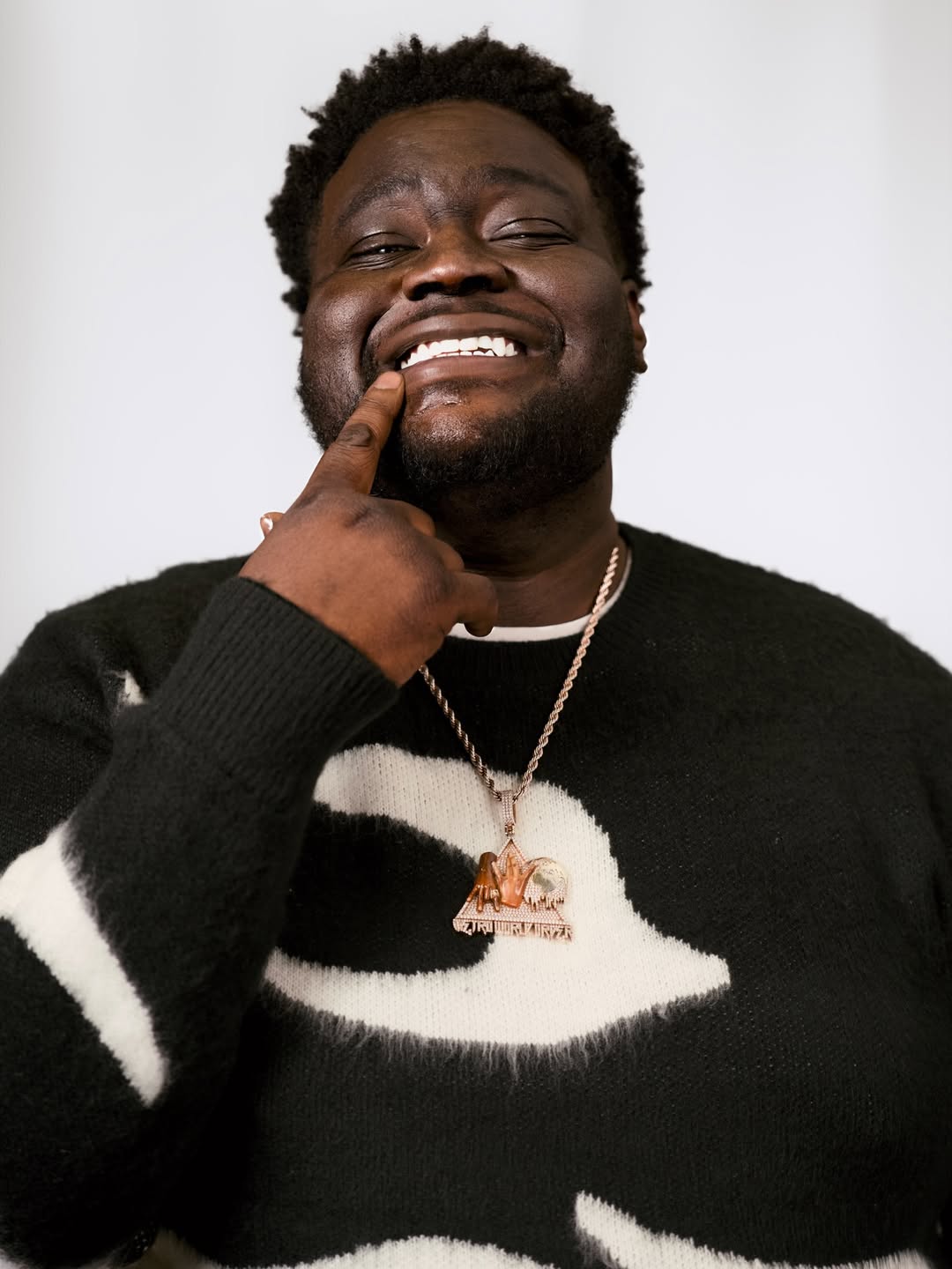 Swayvo Twain touching his lip whilst wearing a black sweater with white patterns and a gold chain necklace